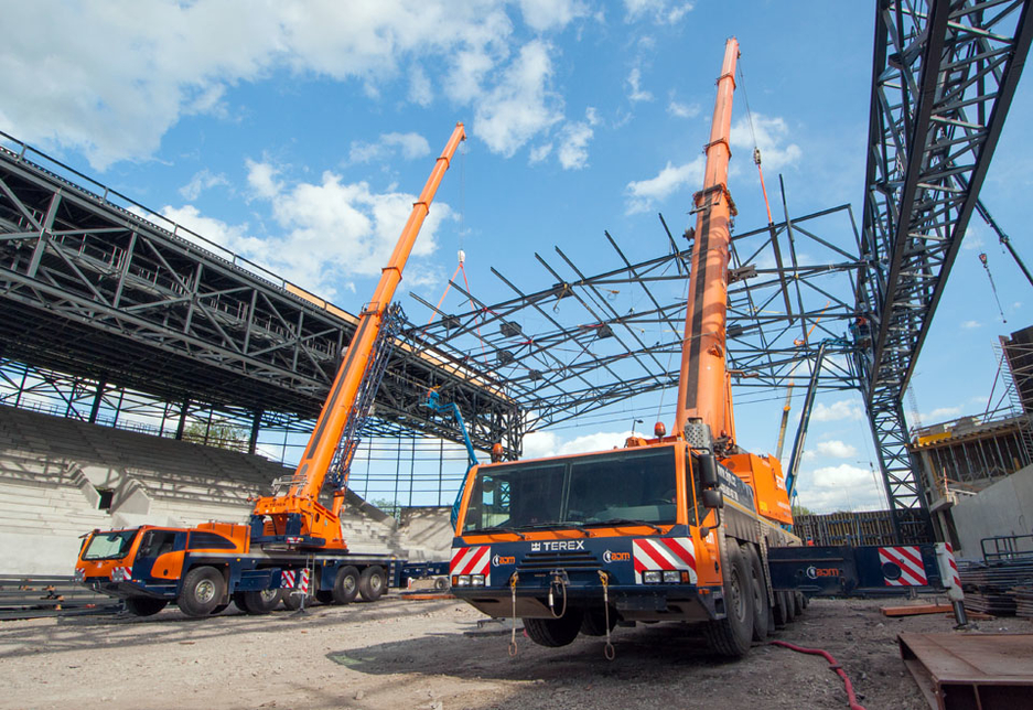A Terex Explorer 5800 and Terex AC 250-1 pair of all-terrain cranes are used to tandem lift pre-fabricated steel roof structures into place during the construction of a sports venue. A Terex Explorer 5800 and Terex AC 250-1 pair of all-terrain cranes are used to tandem lift pre-fabricated steel roof structures into place during the construction of a sports venue.